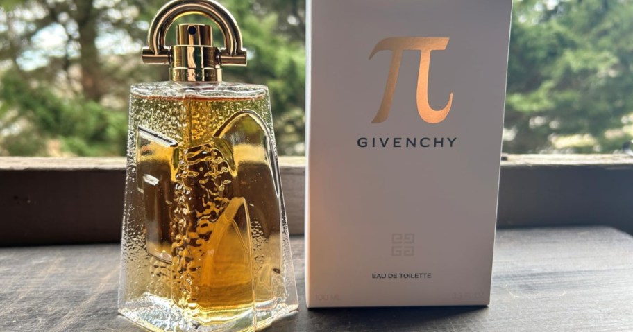A Givenchy Pi perfume bottle with amber liquid and a gold cap stands next to its white box on a wooden surface. Trees are blurred in the background.