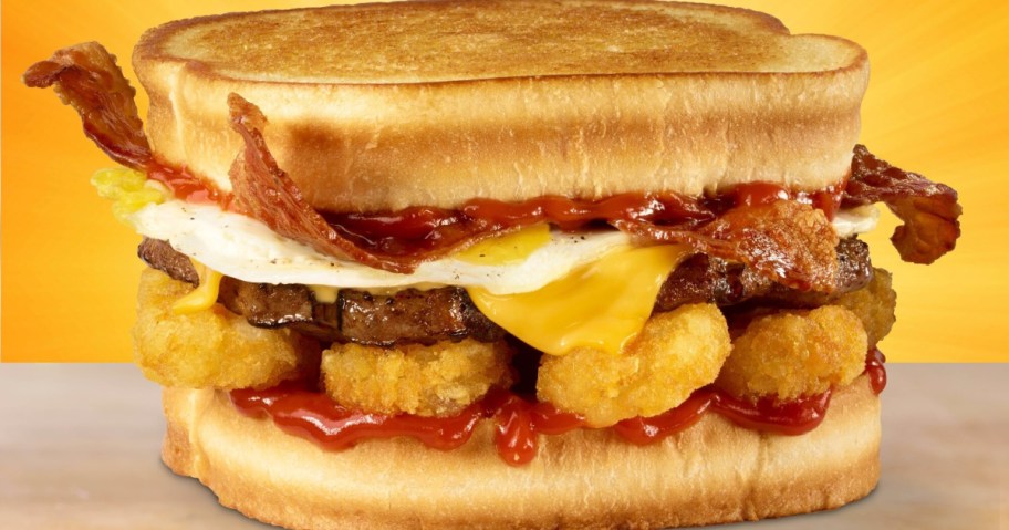 A hearty sandwich with grilled bread, crispy bacon, a sunny-side-up egg, cheese, hash brown rounds, and tangy ketchup, set against an orange background.