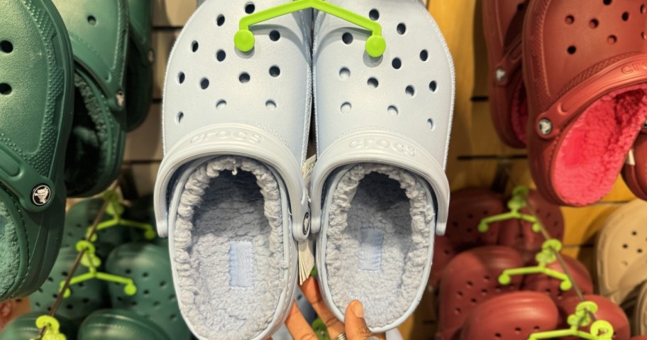 Light blue Crocs with fuzzy lining, hanging on a display. Surrounded by various colored Crocs, a hand holds the pair.