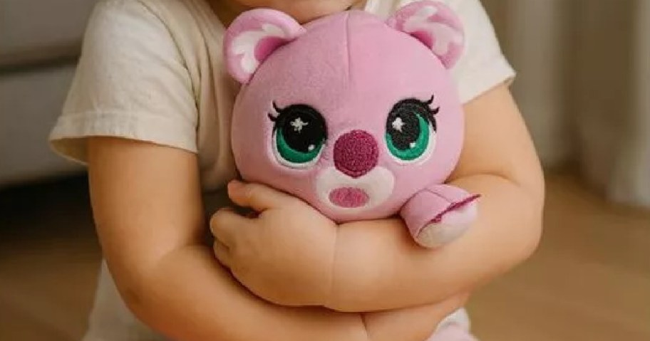 A child in a white shirt hugs a pink Littlest Pet Shop plush with large green eyes and a small round nose.