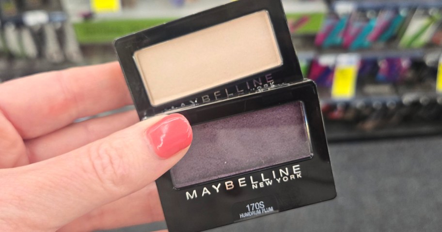 A hand with pink nails holds two Maybelline eyeshadow compacts. The top shade is beige, and the bottom shade is purple. Background shows a blurred store aisle.