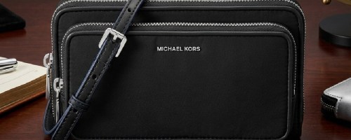 Black Michael Kors crossbody bag with silver zippers on a wooden table. It features an adjustable strap and metallic logo.