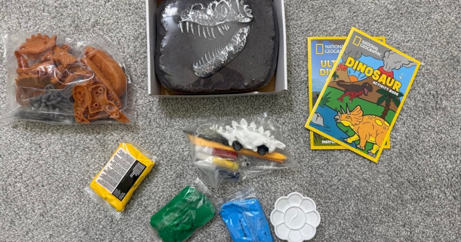 A kids' dinosaur excavation kit on a carpet, featuring a plaster block with a fossil design, plastic dinosaur figurines, two activity booklets, colorful clay, paint set, and a palette.