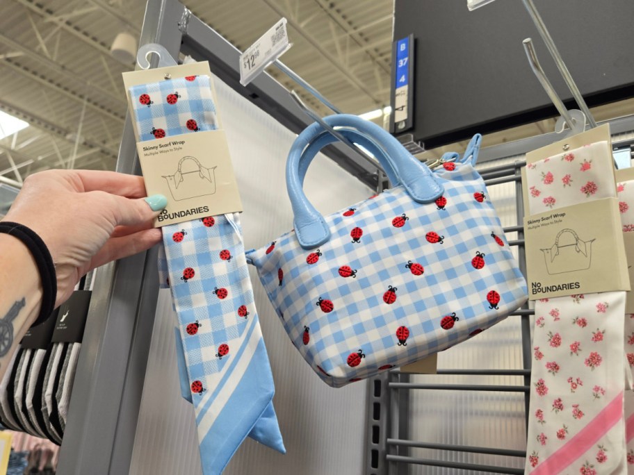 A hand holds a blue checkered scarf with ladybug prints next to a matching bag on a store display.