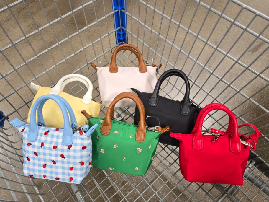 Six colorful handbags are neatly arranged in a shopping cart on a concrete surface. Designs include solid colors, plaid, ladybugs, and floral patterns.