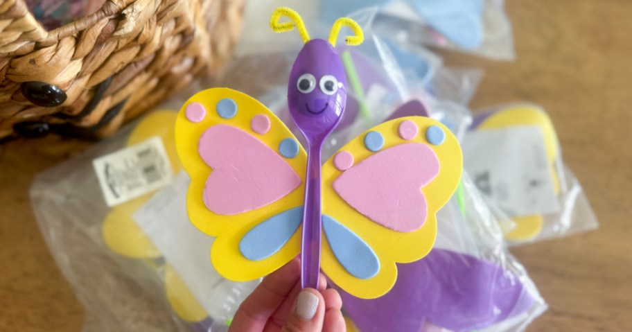 A hand holds a colorful butterfly craft made from a purple plastic spoon, with yellow wings.