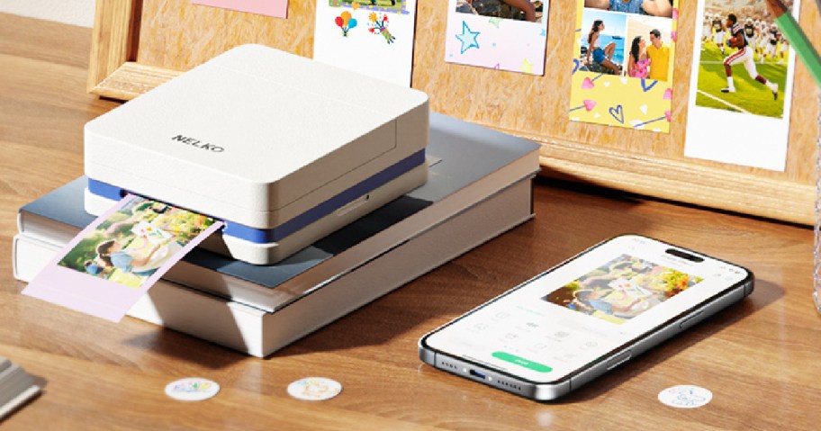 A compact photo printer is printing a colorful image, placed on a stack of books. Nearby, a smartphone displays a matching photo.