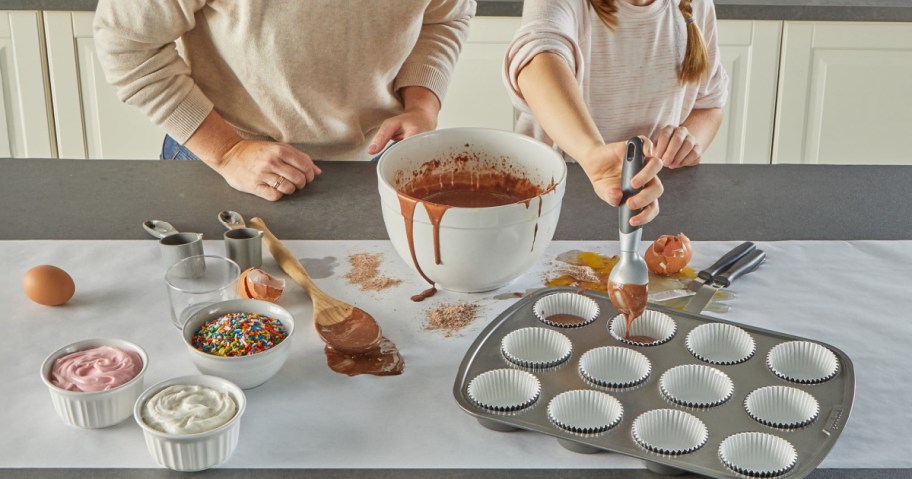 Two people bake cupcakes on top of some Reynolds countertop prep paper, with one scooping chocolate batter into a lined muffin tray. Ingredients like sprinkles and icing are on the counter.