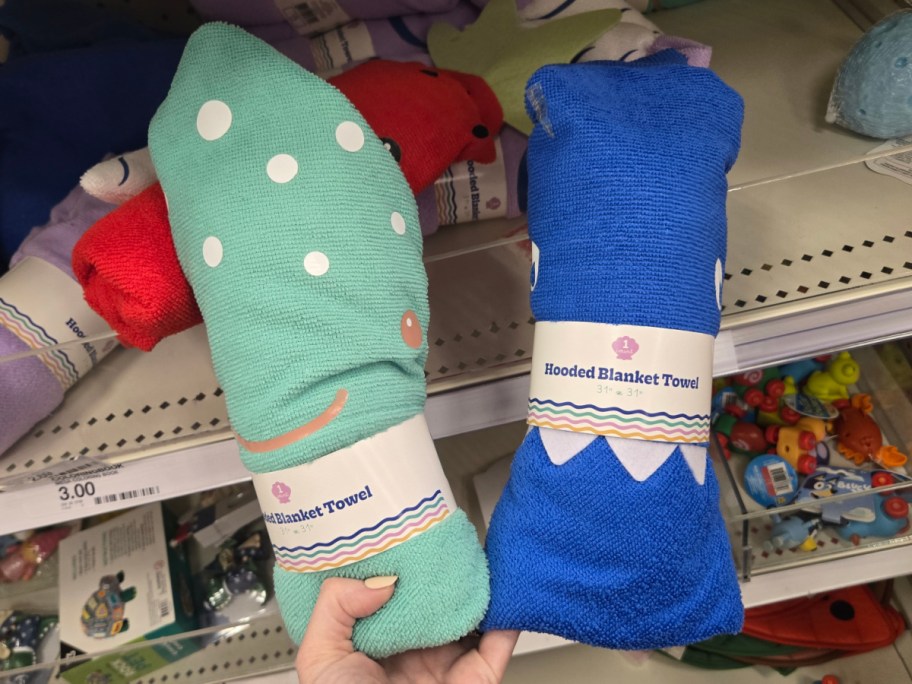 Two hooded blanket towels are displayed in a store. One is green with white polka dots, and the other is blue with a zigzag pattern. Price tags are visible.
