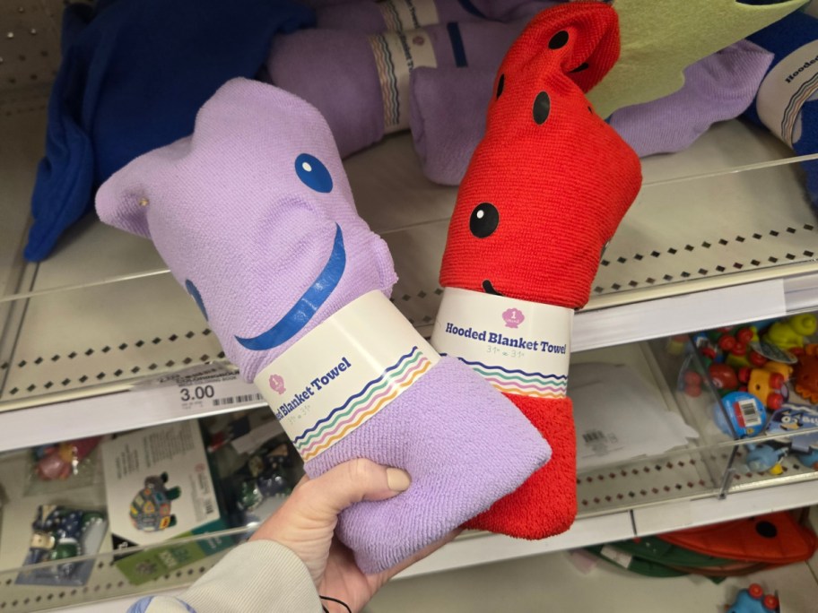 A hand holds two hooded blanket towels in a store. One is purple with a smiling face, and the other is red with black dots.