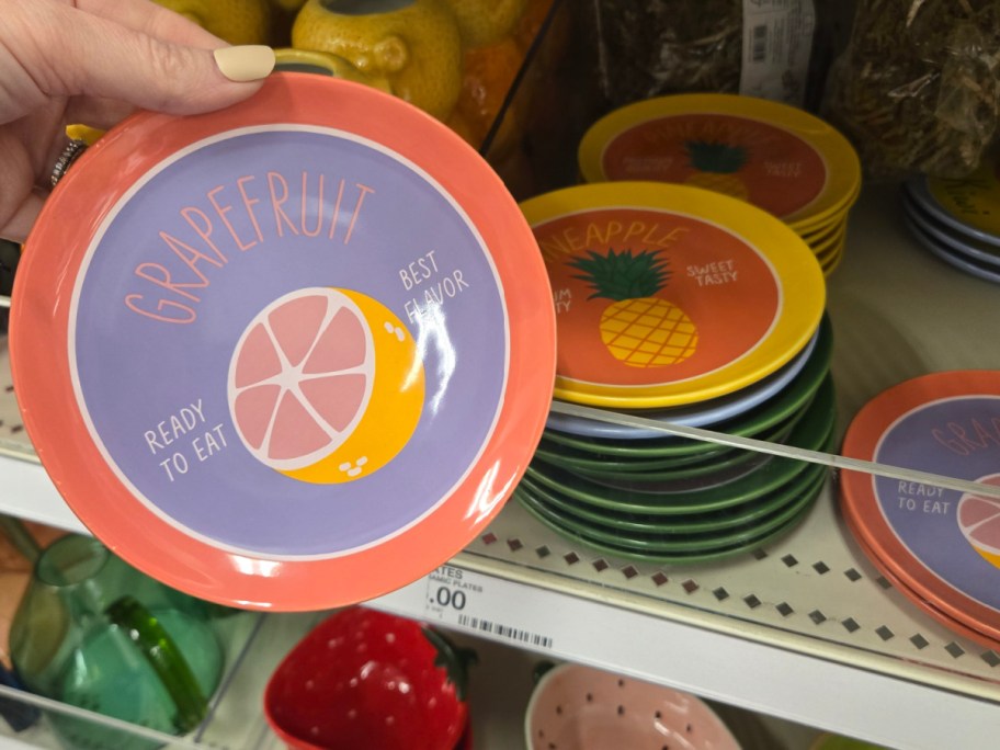 A hand holds a colorful plate with a grapefruit design and the text "Ready to Eat, Best Flavor." Nearby, other plates feature pineapple designs.