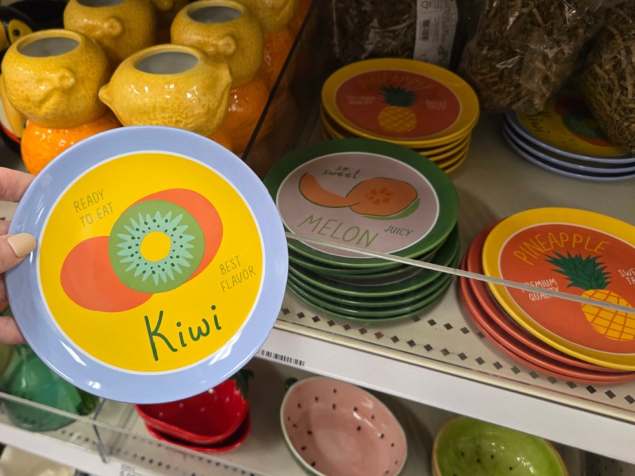 Colorful ceramic plates with fruit designs are displayed on a store shelf. A hand holds a kiwi-themed plate with "Ready to Eat" and "Best Flavor" text.