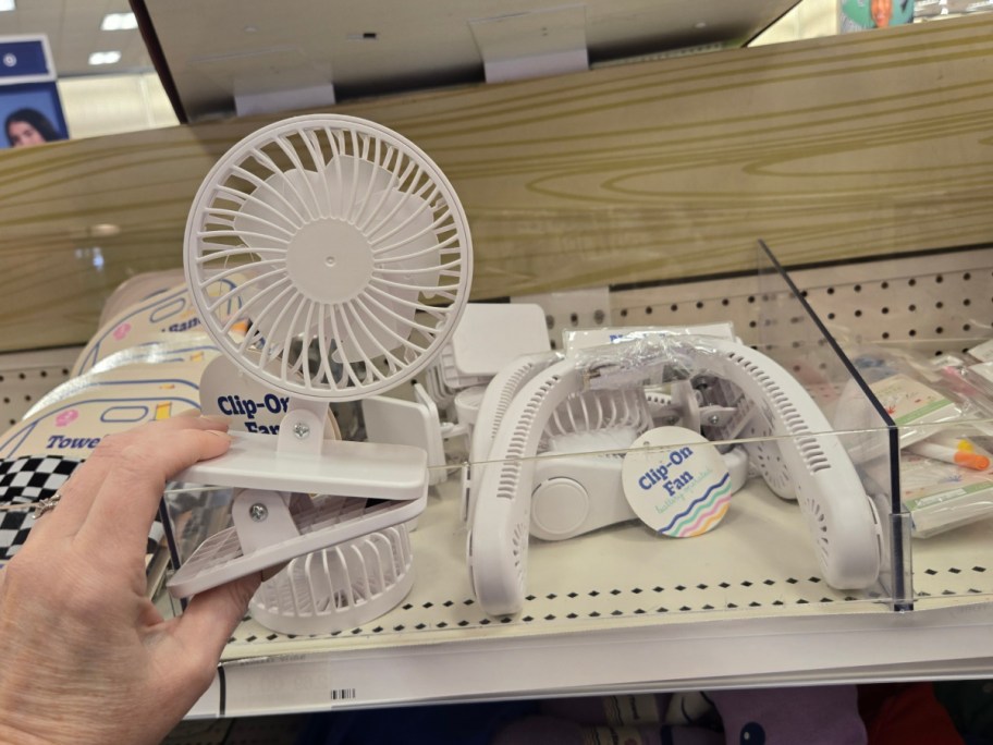 A hand holds a small white clip-on fan in a store aisle. Other similar fans are on the shelf, labeled "Clip-On Fan."
