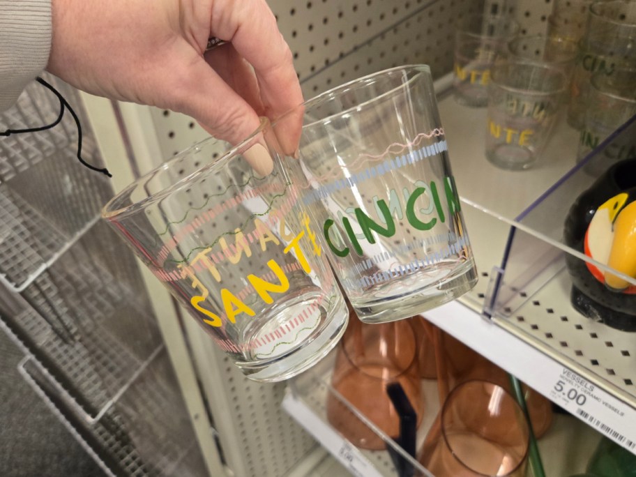 A hand holds two clear glasses with colorful, festive text reading "SANTE" and "CIN CIN," displayed on store shelves with similar glasses and items.