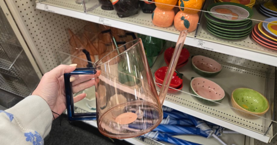 A person holds a transparent pink watering can with a blue handle in a store. Shelves display colorful bowls and decorative items in the background.