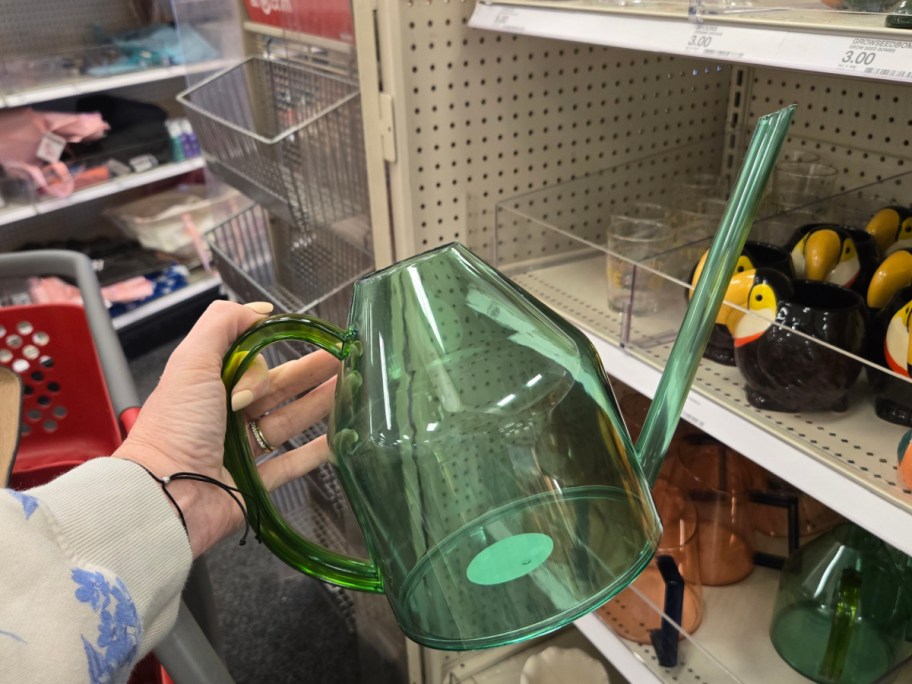 A person holds a translucent green watering can in a store aisle. The shelves display various ceramic mugs, including some with bird designs.