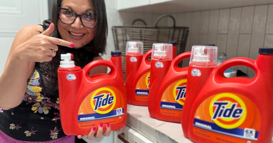 Smiling woman with glasses points to an orange Tide detergent bottle in her hand. Four more bottles are on a counter.