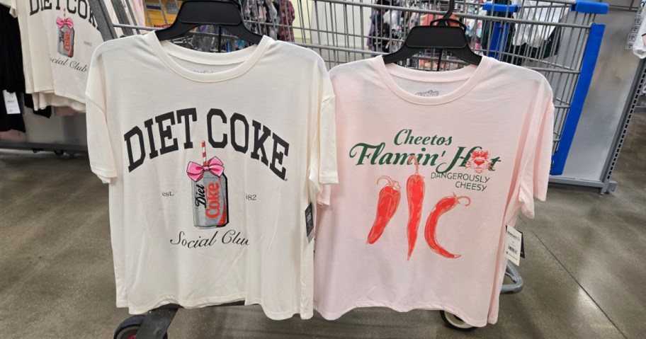 Two graphic T-shirts hanging side by side in a store. The left shirt features a Diet Coke can with a pink bow and text "Diet Coke Social Club." The right shirt shows red chili peppers with the text "Cheetos Flamin' Hot, Dangerously Cheesy."