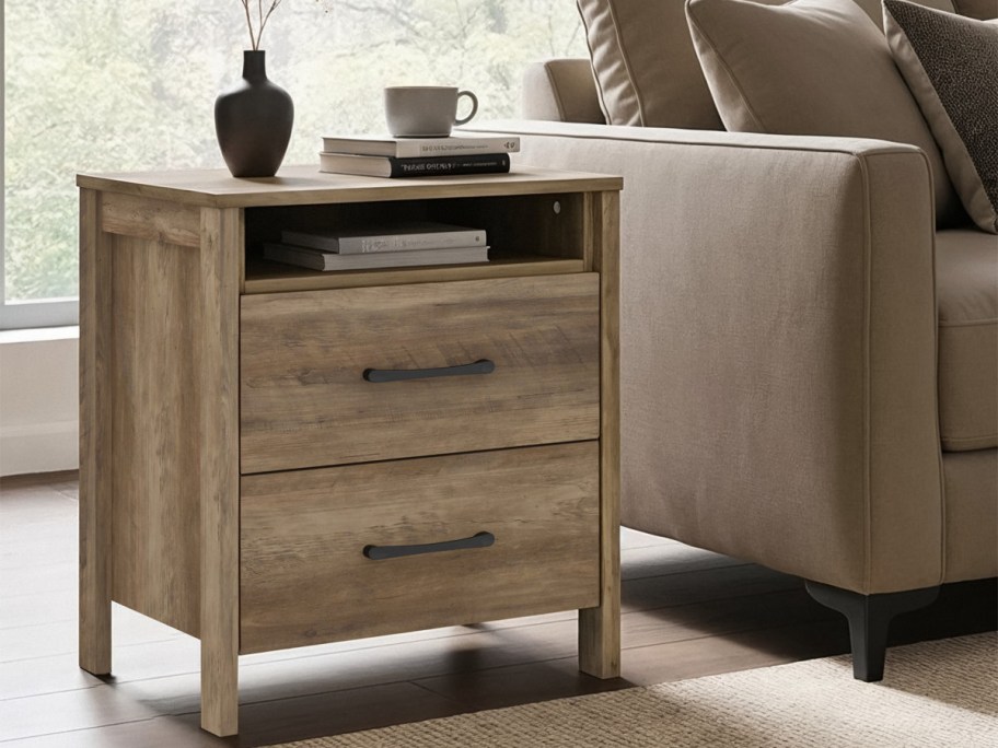 A rustic wooden side table with two drawers and a shelf sits next to a beige sofa. A mug and books rest on top,
