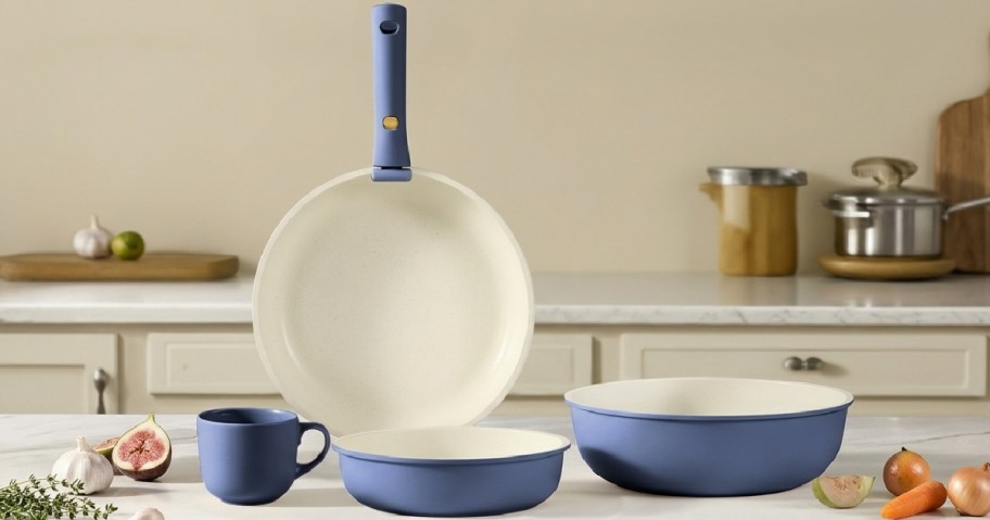 A stylish kitchen set includes a blue frying pan, mug, and two bowls on a marble countertop.