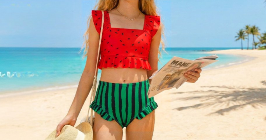 girl wearing watermelon two-piece swimsuit with purse and newspaper on beach with ocean in the background