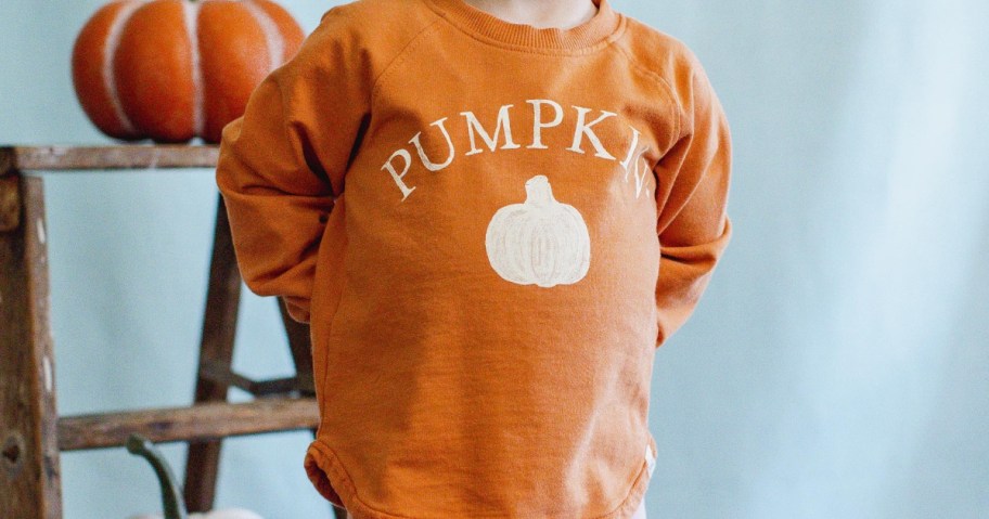 A toddler wearing an orange Burt's Bees Baby organic cotton sweatshirt with a white pumpkin graphic and "PUMPKIN" text.