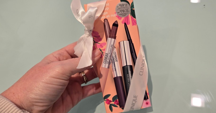 A hand holds a Clinique makeup gift set adorned with a white ribbon. The box features floral designs and showcases three cosmetic products on the cover.
