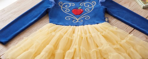 A blue long-sleeve Disney Princess tutu dress featuring a red apple and gold scroll embroidery on the bodice, ruffled tulle sleeves, and a tiered yellow tulle skirt resting on a wooden table.