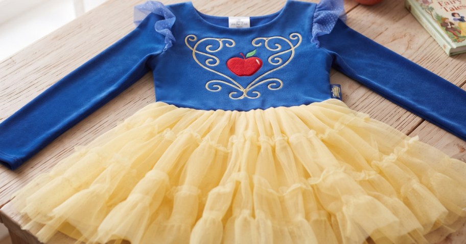 A blue long-sleeve Disney Princess tutu dress featuring a red apple and gold scroll embroidery on the bodice, ruffled tulle sleeves, and a tiered yellow tulle skirt resting on a wooden table.