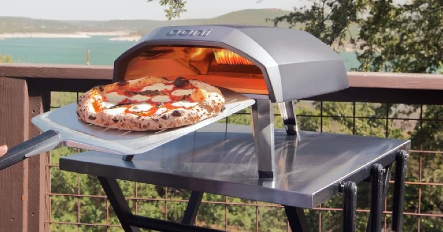 A pizza oven on a patio table with a scenic lake view. A hand uses a paddle to insert a pizza with melted cheese and pepperoni into the oven.