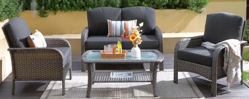 Outdoor patio setting with wicker furniture: a loveseat, two chairs, and a glass-top table. Decor includes cushions, a tray with drinks, and sunflowers.
