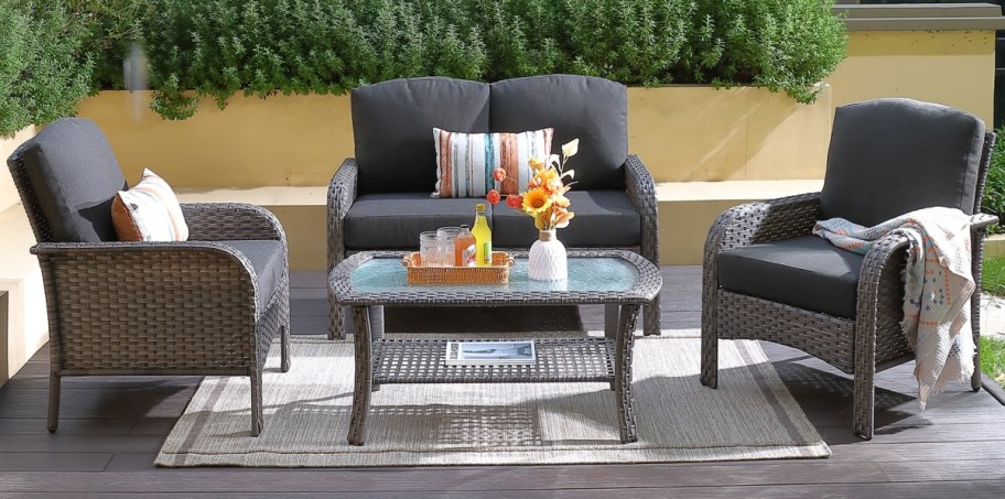 Outdoor patio setting with wicker furniture: a loveseat, two chairs, and a glass-top table. Decor includes cushions, a tray with drinks, and sunflowers.
