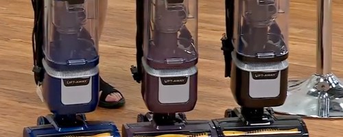 Three Shark vacuum cleaners in blue, purple, and black on a wooden floor.