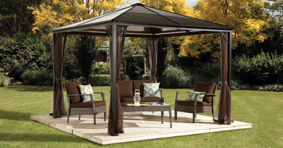 A cozy outdoor gazebo on a wooden deck features brown wicker furniture with cushions, a glass-top table, and is surrounded by lush green foliage.