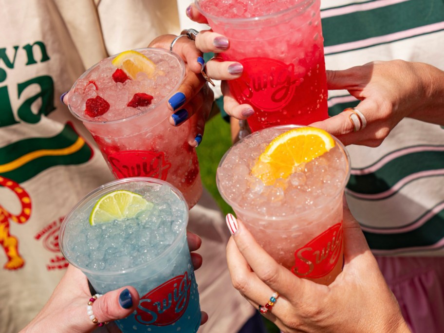 Hands holding four different swig, energy drinks filled with ice and different fruit toppings