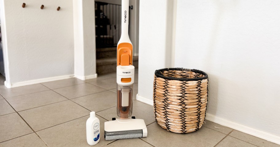 A sleek white and orange vacuum cleaner stands on a tiled floor beside a woven basket and cleaning solution.