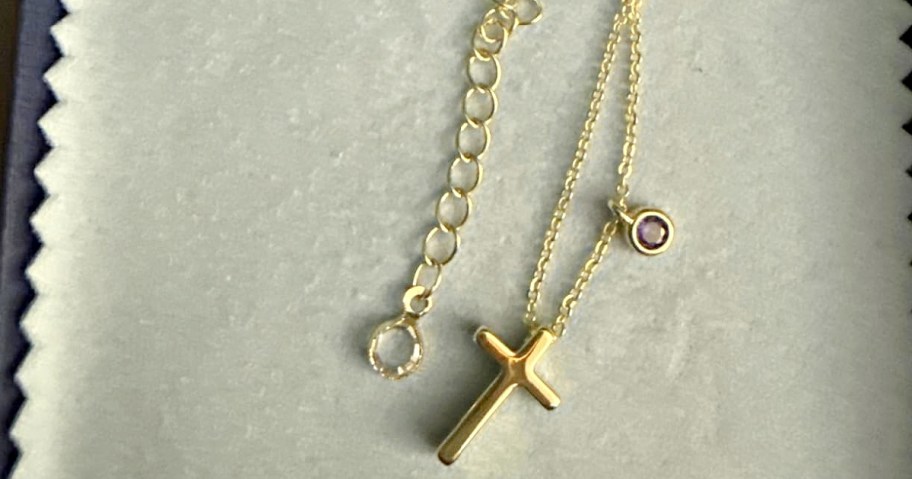 A delicate gold chain necklace featuring a smooth cross pendant and a small round purple gemstone.