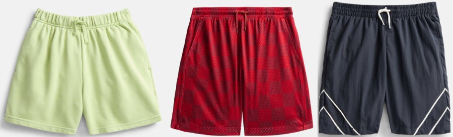 Three pairs of shorts featuring light green sweatshorts, red buffalo plaid athletic shorts with a drawstring, and dark gray athletic shorts with white diagonal stripes.