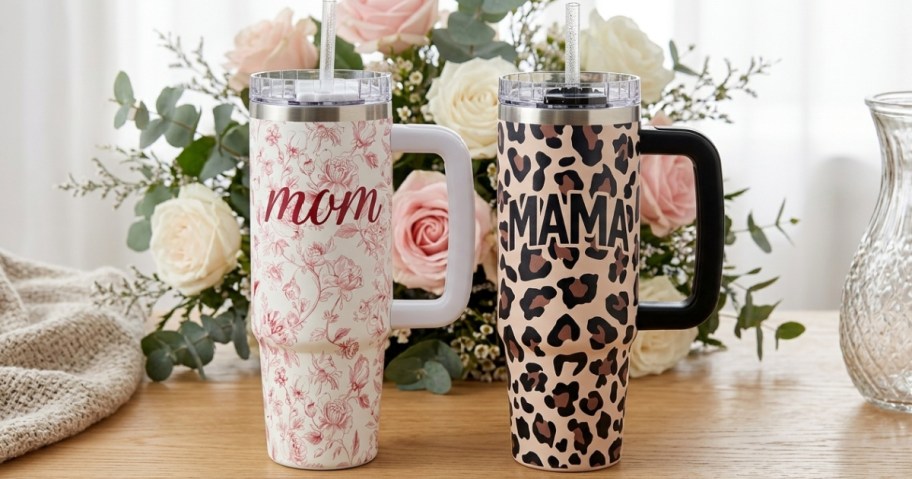 Two tall mugs with handles sit on a wooden table. One features red floral patterns with "mom," and the other has a leopard print with "mama."