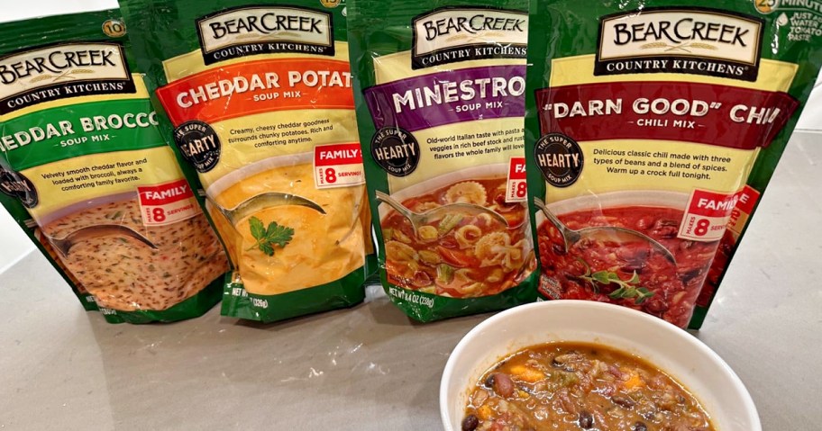 Packets of Bear Creek soup mixes, including Cheddar Broccoli, Cheddar Potato, Minestrone, and Chili, are displayed behind a bowl of mixed soup.