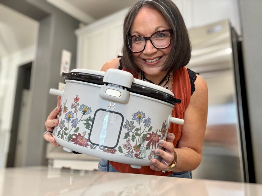 A smiling woman holds a floral-patterned slow cooker in a bright kitchen. The background includes white cabinets.