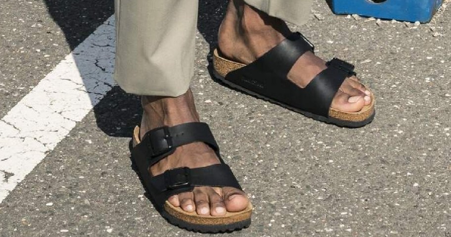 Feet in black two-strap sandals with buckles, worn on a sunlit, textured asphalt road.