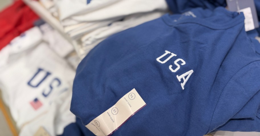 A stack of folded t-shirts with "USA" printed on the front in white letters. The shirts are in various colors, including blue and white.