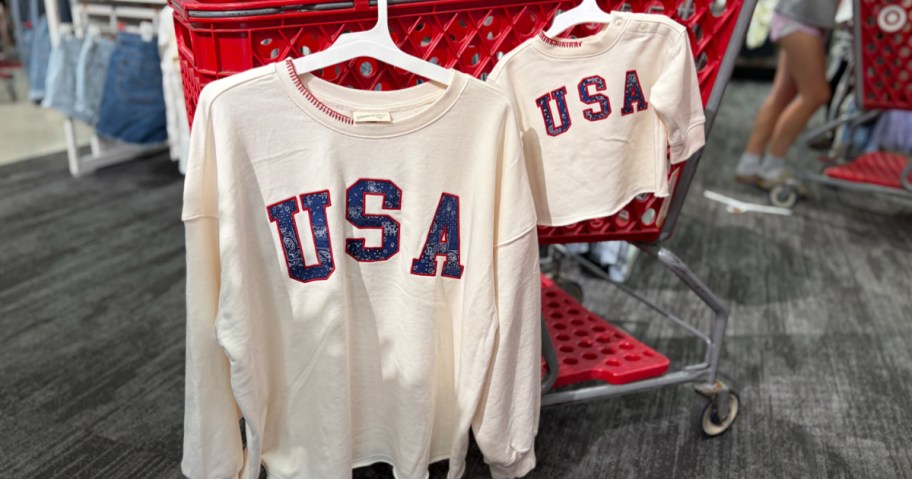 Two clothing items featuring "USA" printed in red and blue, displayed on hangers in a shopping cart.