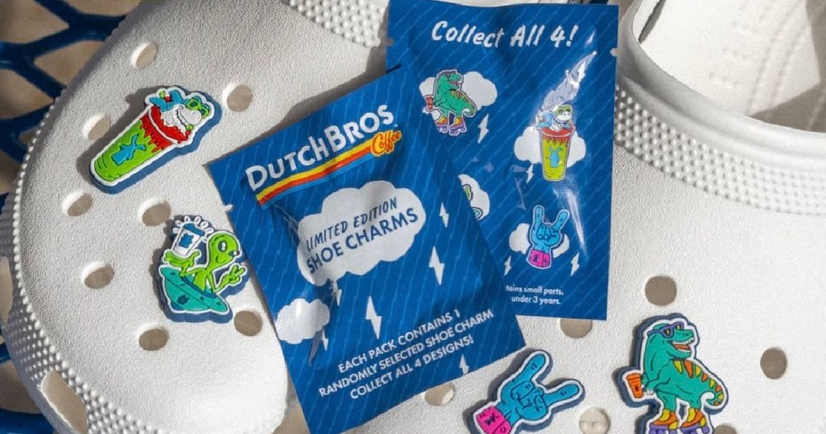 White shoe with colorful Dutch Bros limited edition shoe charms, featuring dinosaurs and drink designs. Blue packaging notes "Collect All 4!"