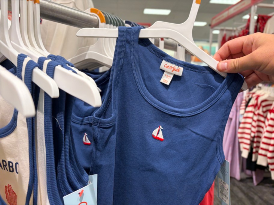 A person holding a blue tank top with a small sailboat design, hanging on a rack in a store.