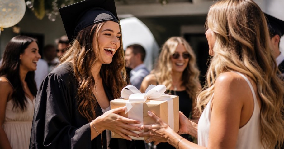 girl wearing graduation cap and gown holding gift from woman at party