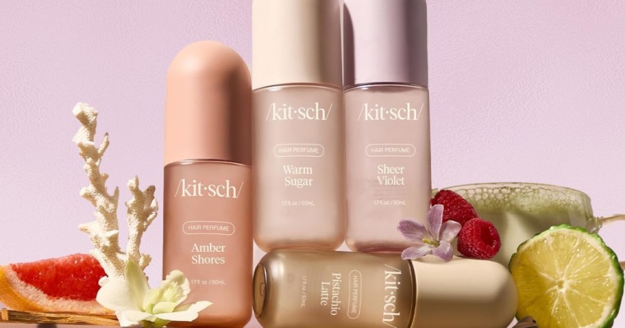 Four pastel-colored bottles of Kitsch hair perfumes are surrounded by grapefruit, lime, raspberry, and coral accents, set against a soft pink background.