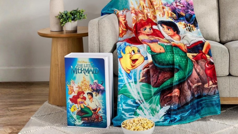 little mermaid throw and vhs box on a sofa with bowl of popcorn