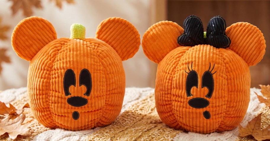 Two orange corduroy pumpkin plushies shaped like Mickey and Minnie Mouse heads sitting on a woven blanket surrounded by autumn leaves.